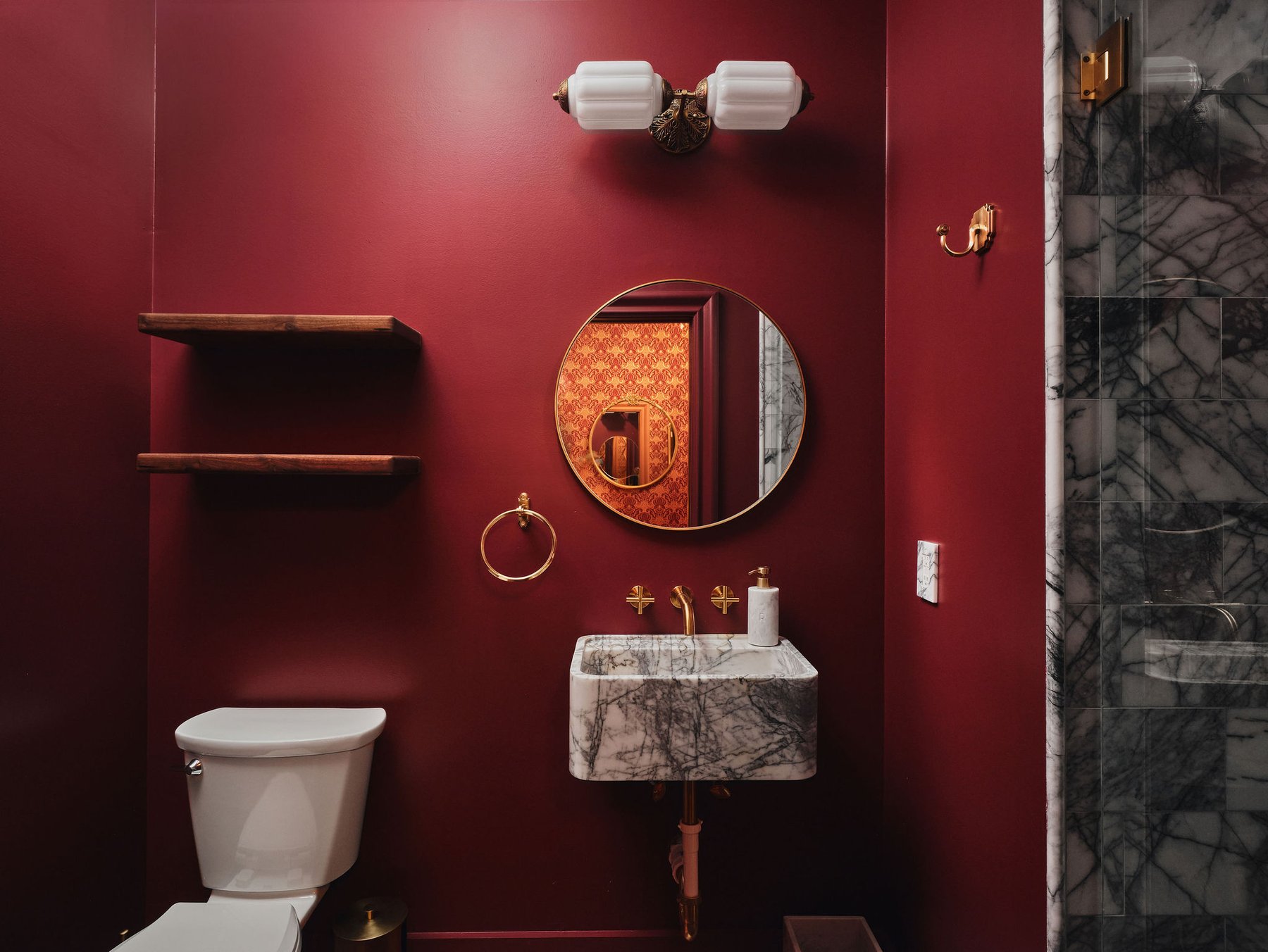 Powder room in deep wine lacquer with a marble basin, brass fittings and a round mirror carrying the damask dressing room beyond